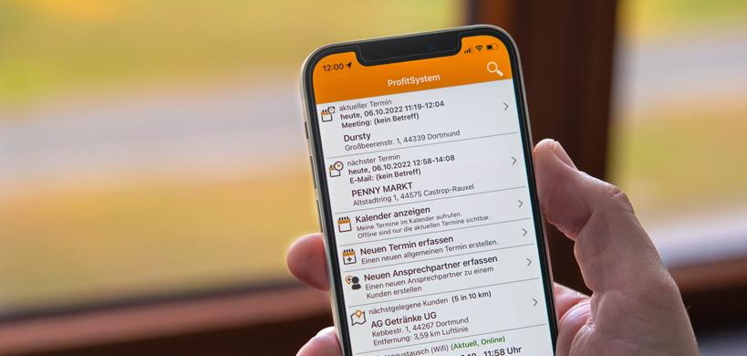 Eine Person hält ein Smartphone, auf dem eine CRM-App mit dem Namen "ProfitSystem" geöffnet ist. Der Bildschirm zeigt eine Liste von Terminen und Aufgaben. Im Hintergrund sind unscharfe Fenster und Natur zu sehen.