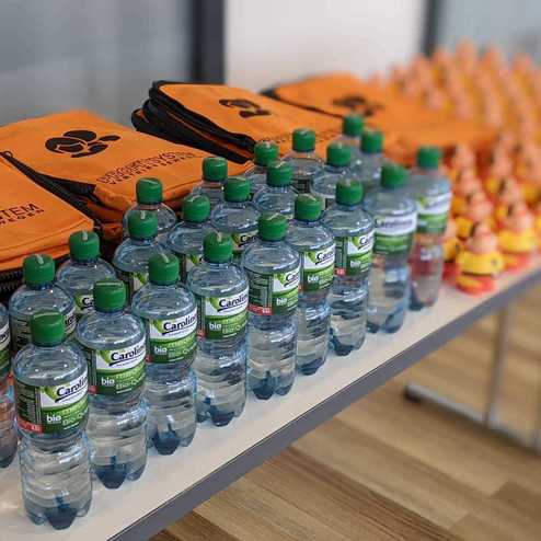 Orangefarbene Taschen mit einem Logo und Wasserflaschen von Carolinen auf einem Tisch, daneben kleine gelbe Spielzeugenten.